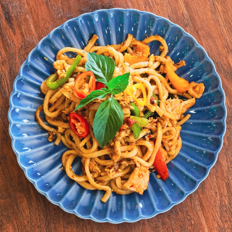 Udon noodles with chicken, colorful bell peppers, and basil on a blue scalloped plate.