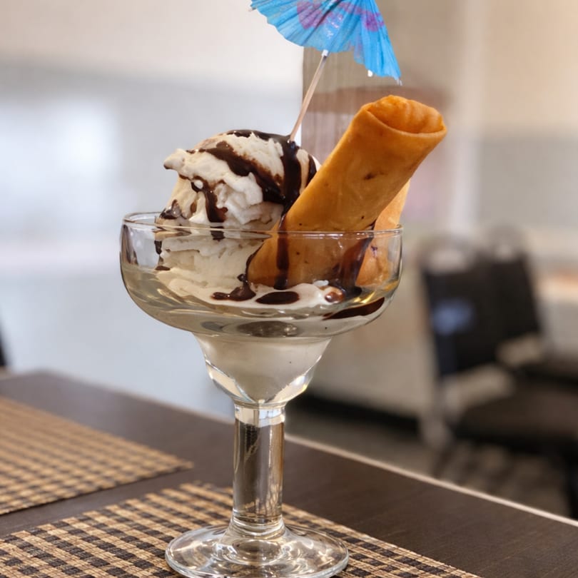 Vanilla ice cream sundae with chocolate syrup, a fried spring roll, and a blue umbrella.