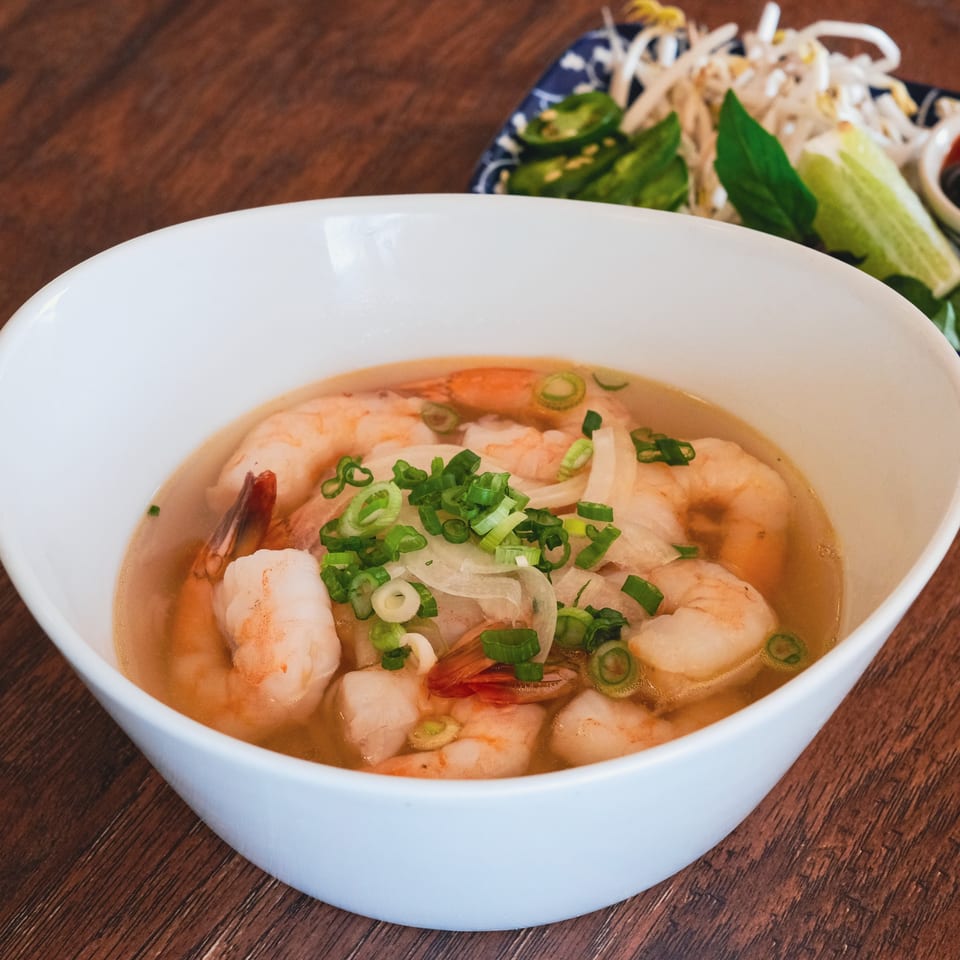 Shrimp pho in a white bowl, topped with green onions and white onions, with a side of bean sprouts.