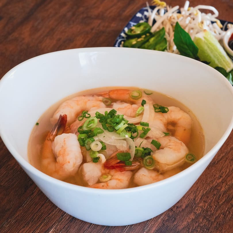 Shrimp pho in a white bowl, topped with green onions and white onions, with a side of bean sprouts.