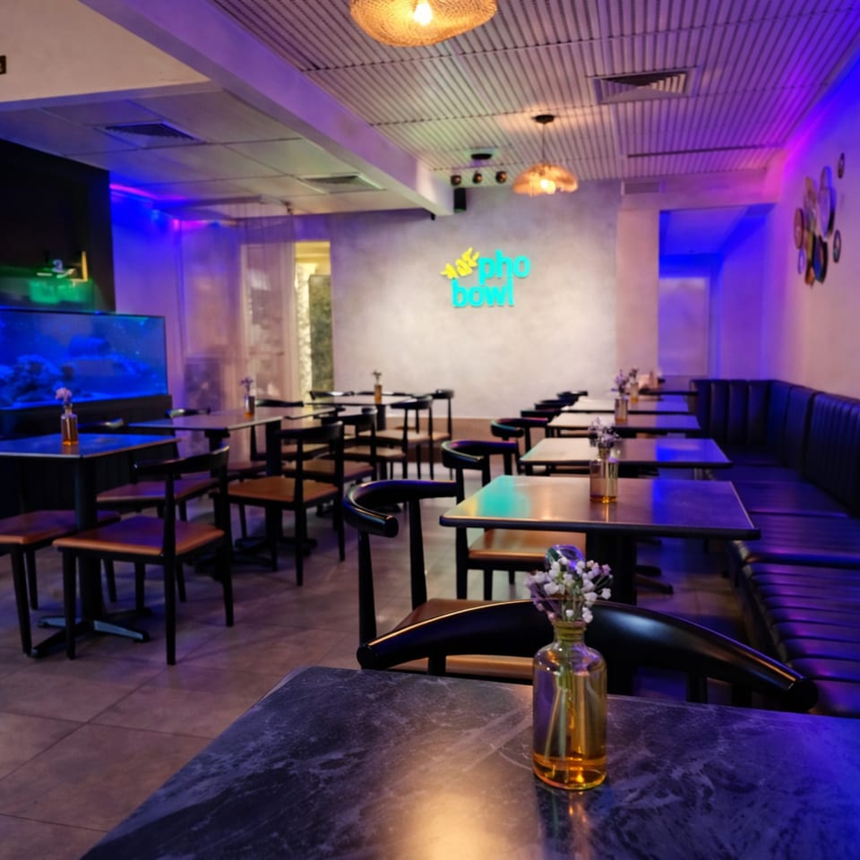 A modern restaurant interior with blue and purple lighting, tables, chairs, and a "pho bowl" neon sign.