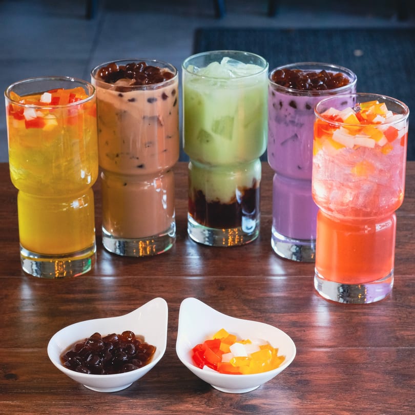 Five colorful bubble tea drinks and two small bowls of toppings on a wooden table.