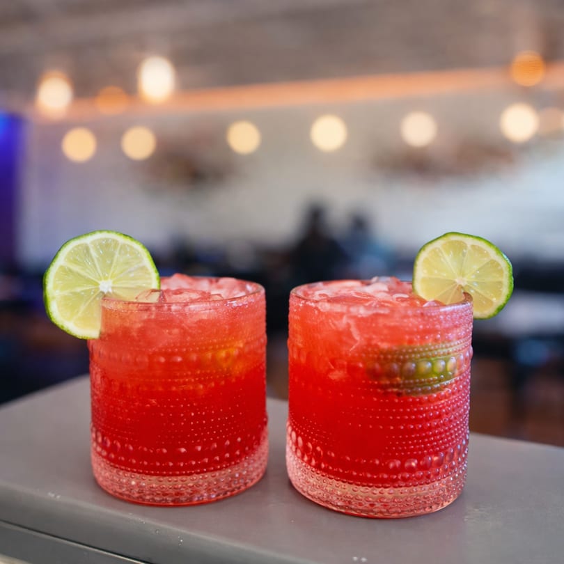 Two vibrant red cocktails with lime garnishes sit on a bar, with blurred lights in the background.