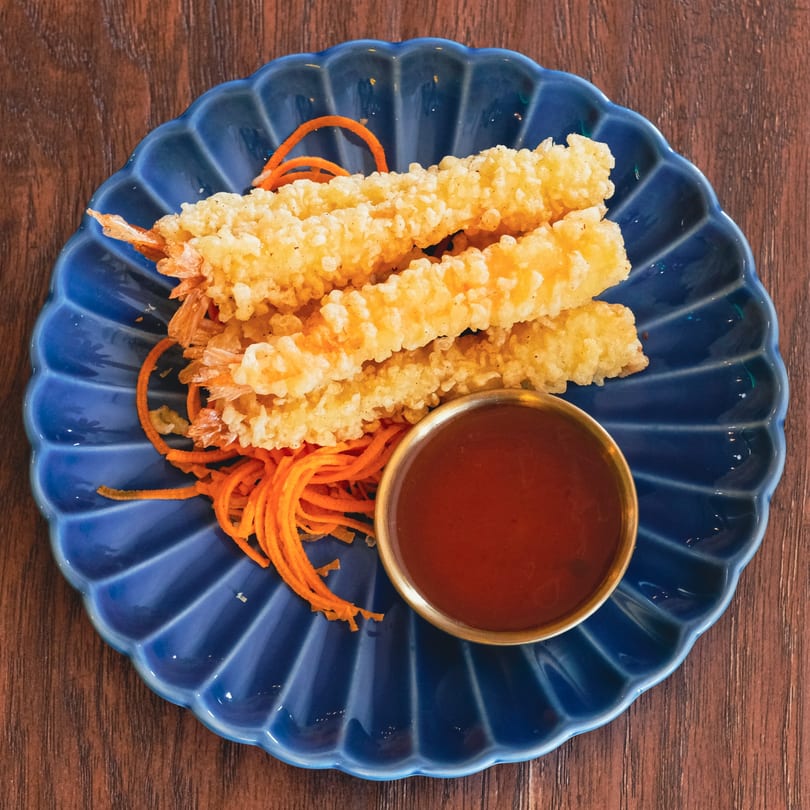 Crispy fried shrimp tempura with shredded carrots and a side of dipping sauce on a blue plate.