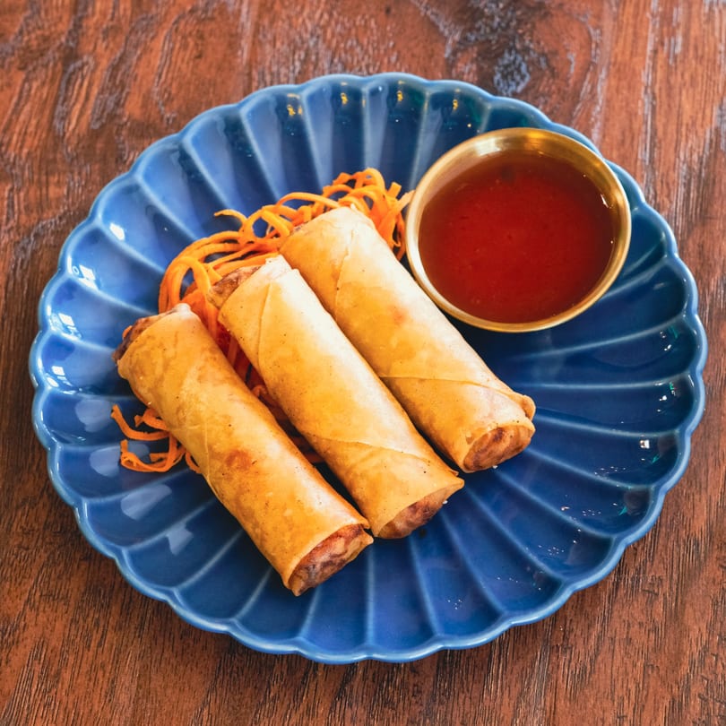 Three golden spring rolls with shredded carrots and a small bowl of red dipping sauce on a blue plate.