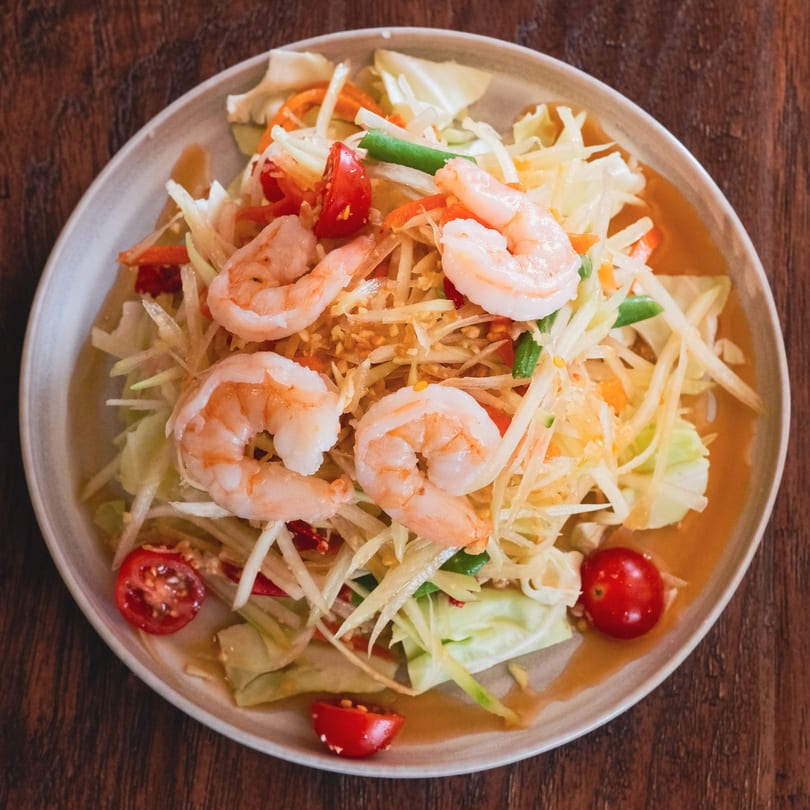 Som Tum, a Thai green papaya salad with shrimp, tomatoes, and cabbage, on a white plate.